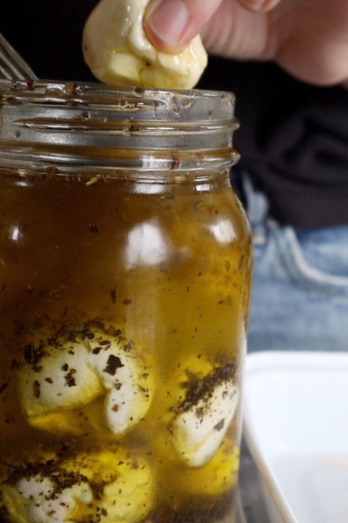 A hand pulling a marinated mozzarella ball out of a jar of olive oil 